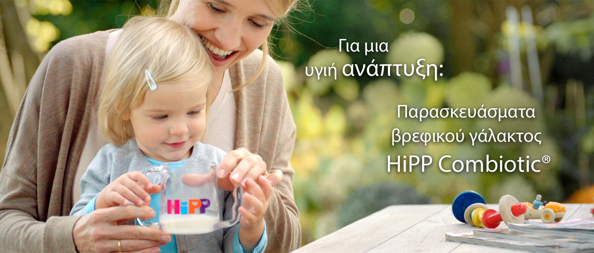 Μια γυναίκα και ένα μικρό παιδί βρίσκονται σε εξωτερικό χώρο, με τη γυναίκα να κρατάει ένα ποτήρι με τη λέξη "HiPP" και το παιδί να το κοιτάζει.
