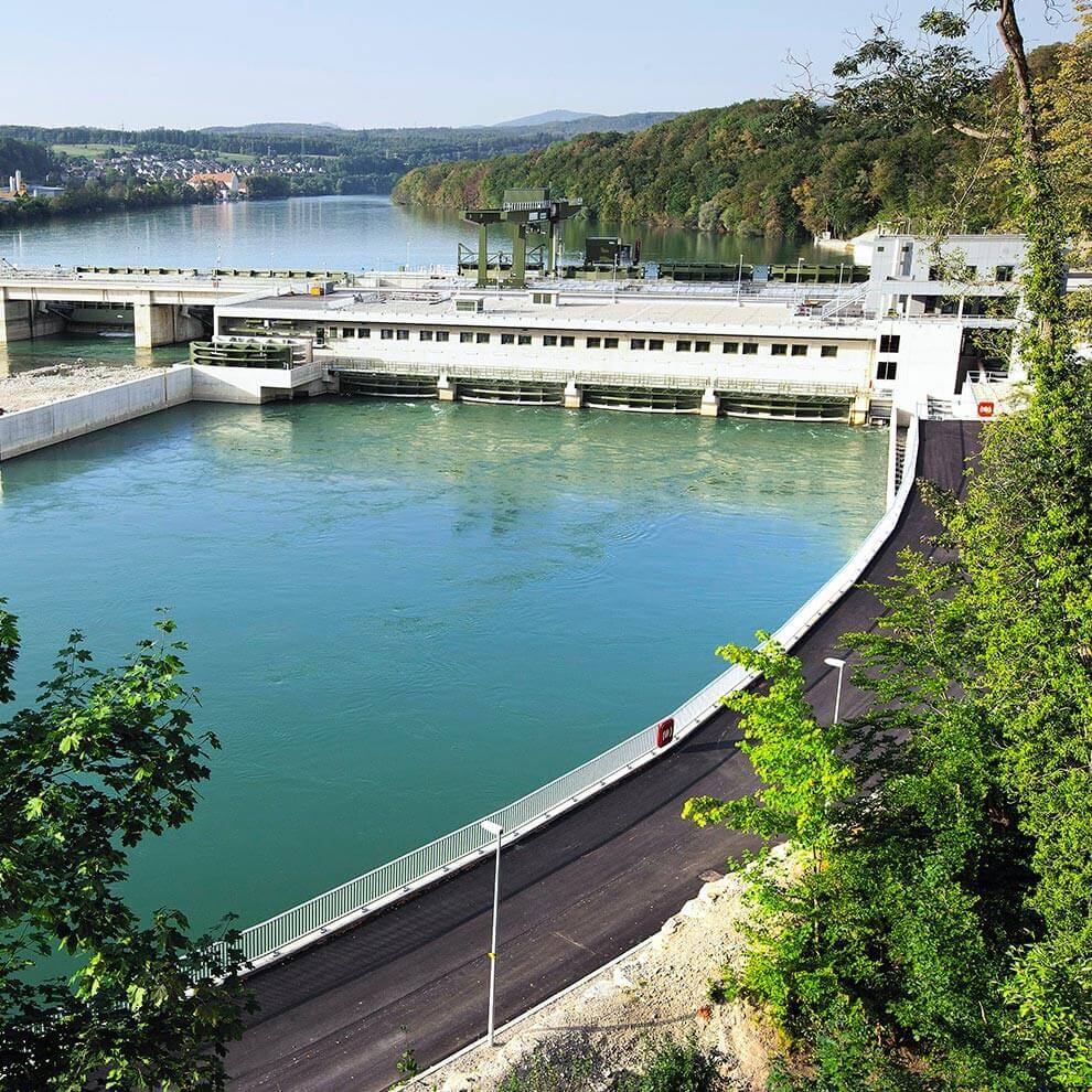 A dam facility with a roadway, surrounded by a river and lush greenery, with a town in the background.