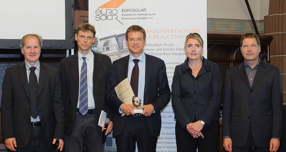 Five individuals stand in front of a promotional banner for 'EUROSOLAR' at an event, with one holding a trophy and a certificate.