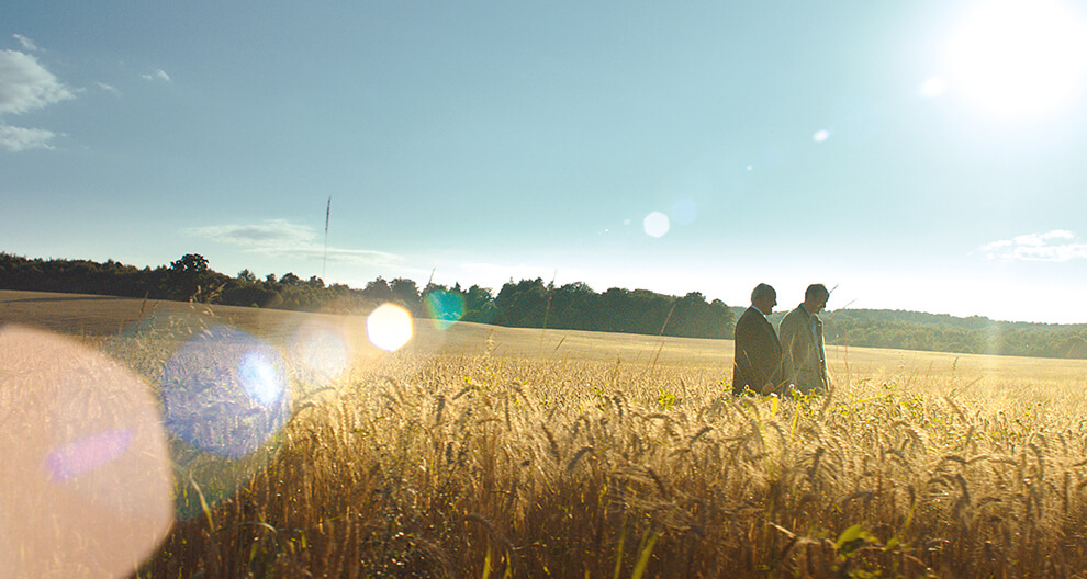Two individuals stand amidst a field of tall grass under a clear sky with a bright sun.