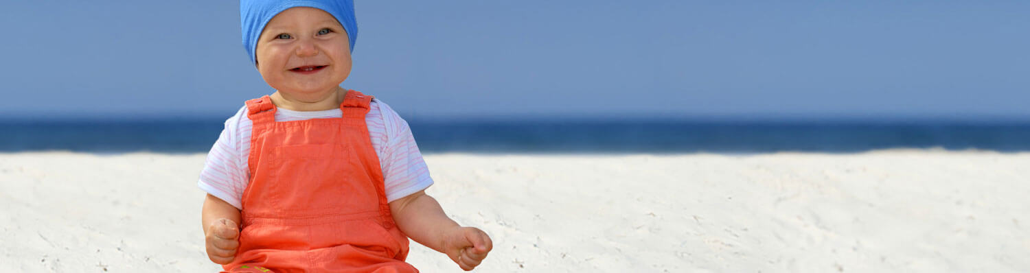 A baby in a blue hat and orange overalls sits on a sandy beach with the ocean in the background.