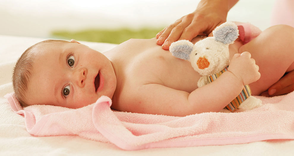 A baby lies on a pink towel, holding a plush toy mouse, with a hand gently resting on the baby's chest.