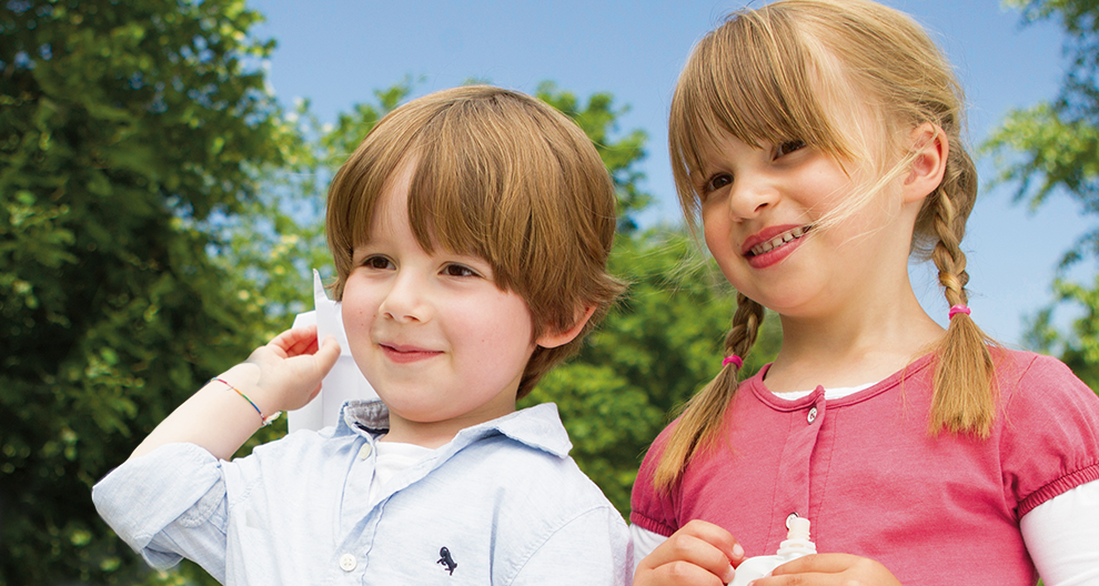Two children, a boy and a girl, are outdoors with trees in the background.