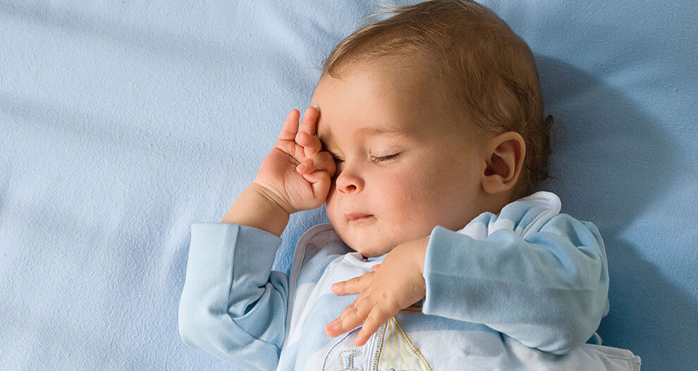 A baby lies on a blue sheet, wearing a light blue outfit with a sailboat design, covering one eye with a hand.