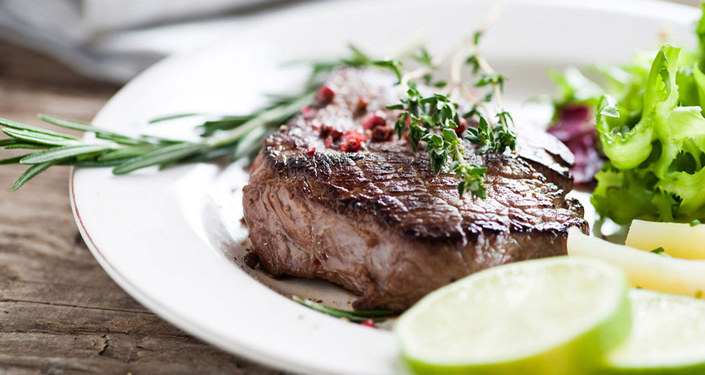 A steak with rosemary and thyme garnish, served with a side of salad and lime slices.