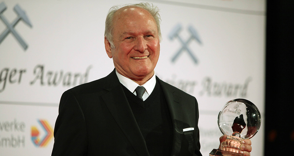An elderly man in a black suit stands on a stage holding a transparent award with a globe design, against a backdrop featuring the words "Schweizer Award" and logos.