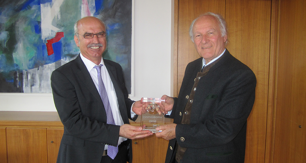 Two men in suits stand in a room with a painting behind them, one presenting a clear award with text to the other.